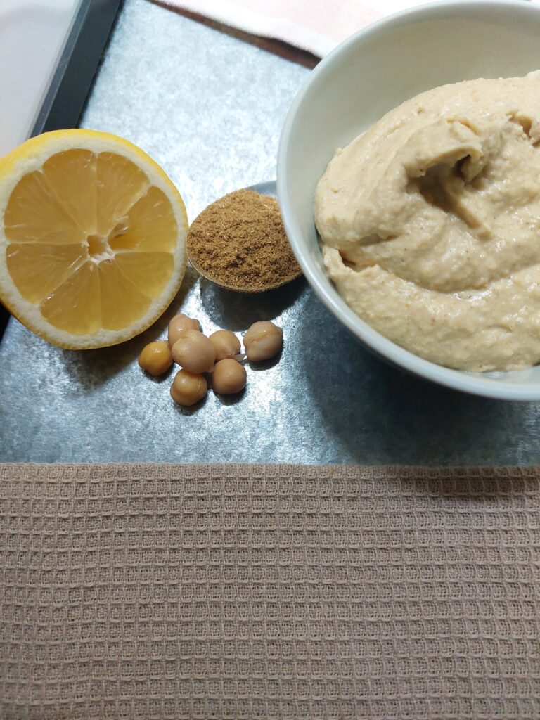 Basic hummus in a bowl next to the main ingredients, chickpeas, cumin and and a lemon cut in half