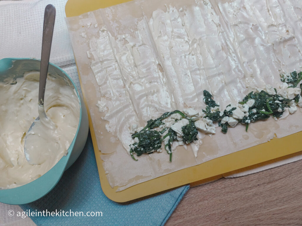 A sheet of filo pastry is laying on top of a yellow cutting board, with a blue napkin peaking out on the sides. The filo pastry is brushed with a mascarpone mixture and a row of blanched spinach and crumbled feta cheese is at the bottom of the filo sheet. To the left is a blue bowl with more mascarpone mixture and a spoon standing up in the bowl.