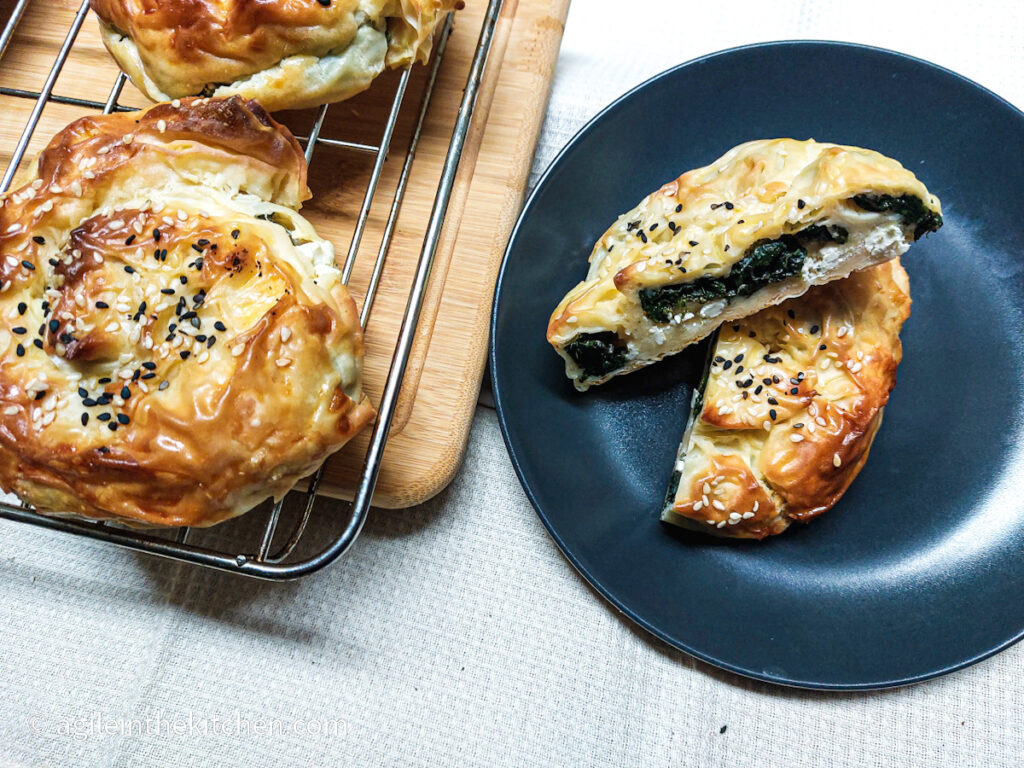 To the left, a Börek straight from the oven is resting on a wire rack, while to the right is a black plate with a Börek but in half is showing the spinach and feta cheese filling.