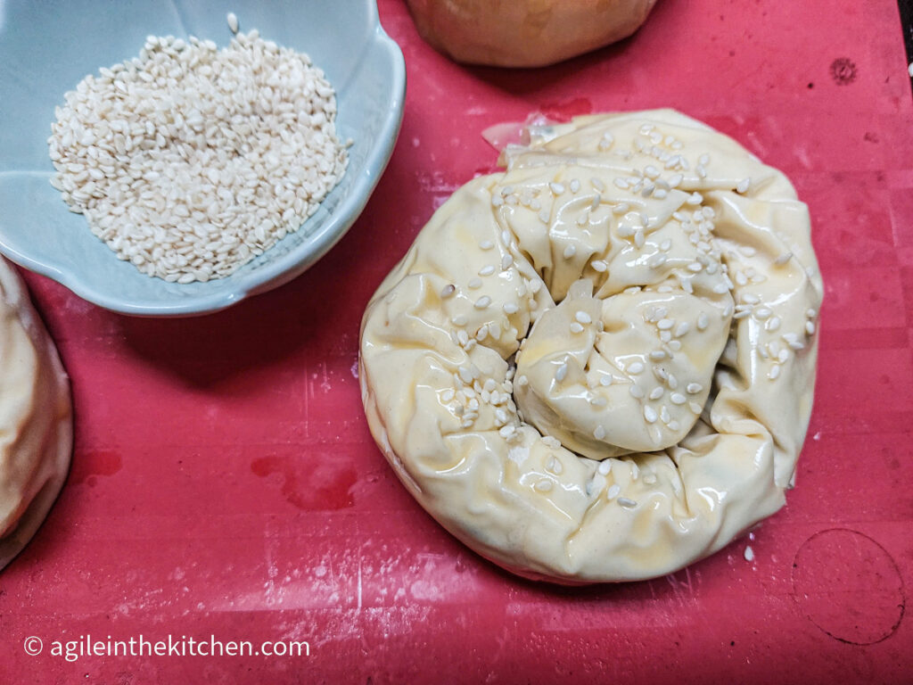 On a red silicone cookie sheet, a rolled up and egg washed Börek is waiting for a sprinkling of white sesame seeds in a blue flower shaped bowl.