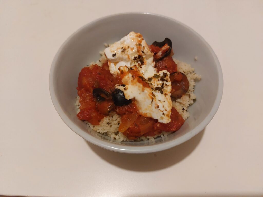 Baked feta cheese in tomato sauce with black olives on a bed of couscous in a white bowl on a white background.