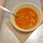 A Moroccan tomato soup with chickpeas in a green bowl on top of a beige napkin and metallic work surface.