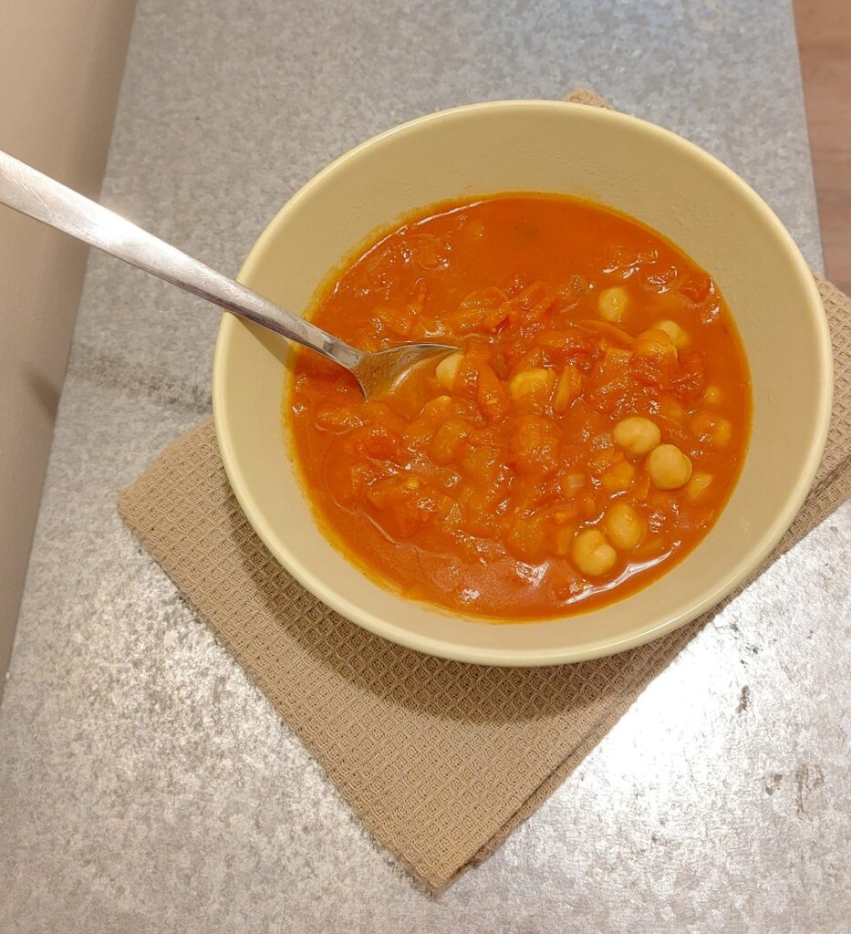 A Moroccan tomato soup with chickpeas in a green bowl on top of a beige napkin and metallic work surface.