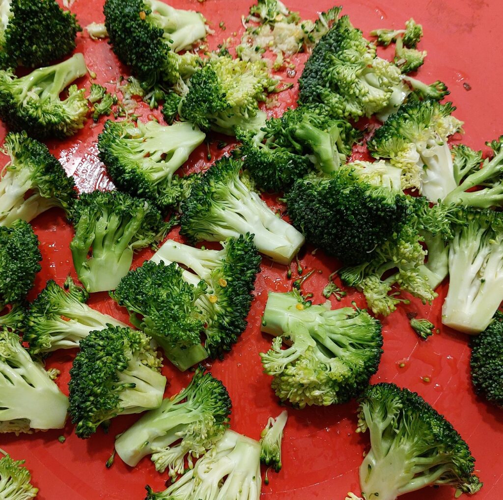 Broccoli florets laid out on a cooking sheet with a red background with olive oil, chili flakes and minced garlic, ready to go into the oven