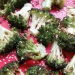 Roasted broccoli florets straight from the oven, sprinkled with white sesame seeds on a red background.