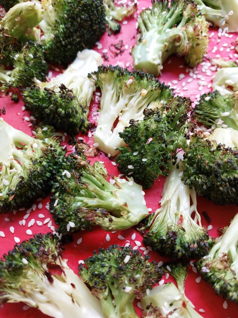 Roasted broccoli florets straight from the oven, sprinkled with white sesame seeds on a red background.