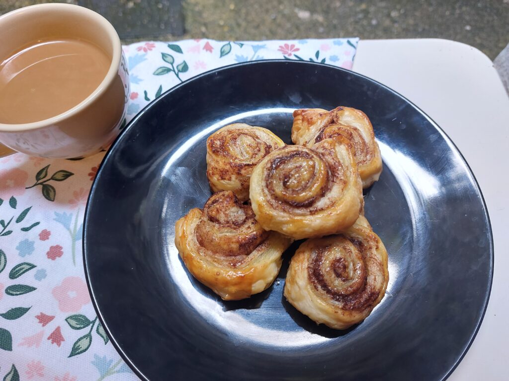Five cinnamon swirls on a black plate, next to a cup of coffee in a beige cup, all sitting on a flowery table cloth. 