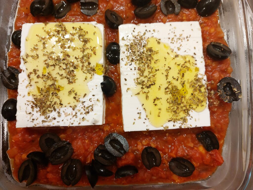 Tomato sauce in a glass dish with black olives and two feta cheese bricks, drizzled with olive oil and sprinkled with oregano.
