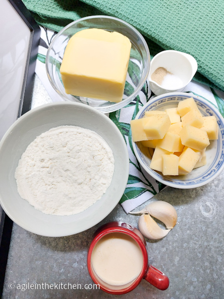 Butter, salt, pepper, cheese, garlic, milk, flour, ingredients laid out to make Mac n' Cheese sauce. Underneath the ingredients a white cloth with green leaves.