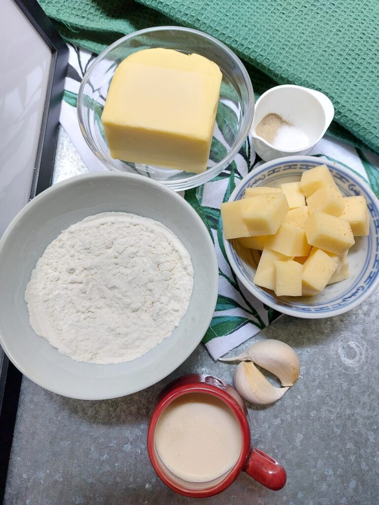 Mac n Cheese ingredients in various 
bowls laid out on a green cloth: butter, salt and pepper, cheese in cubes, two garlic gloves, a cup of milk, flour.