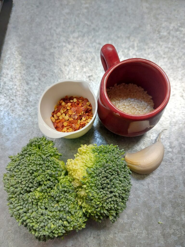 Ingredients for roasted broccoli laid out on a metallic background: Chili flakes, sesame seeds, garlic clove and broccoli florets