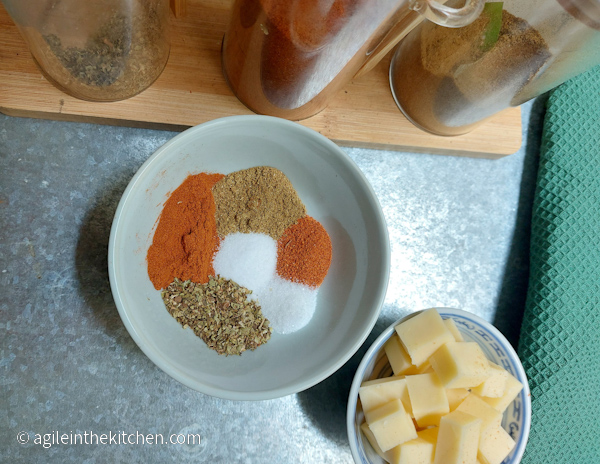 A white bowl with six ingredients for taco seasoning, next to a bowl of cheese cut in cubes. In the background three glass containers with spices, sitting on a wooden plate.