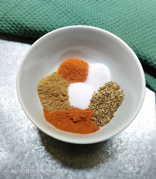 A white porcline bowl with six different spices laid out. The bowl sits on a silver table with a green cloth.