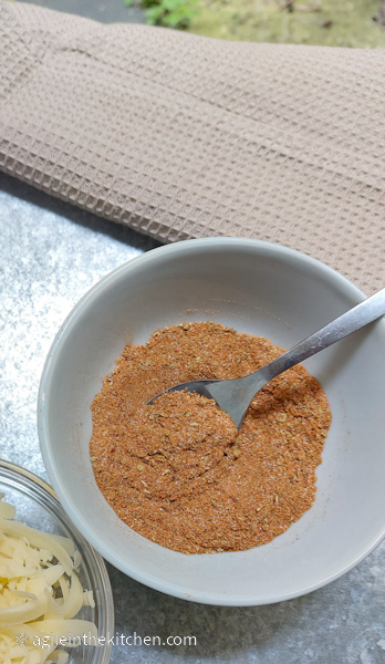 Taco seasoning mixed up in a white bowl and a silver coloured spoon. In left hand corner there is a bowl of shredded cheese.