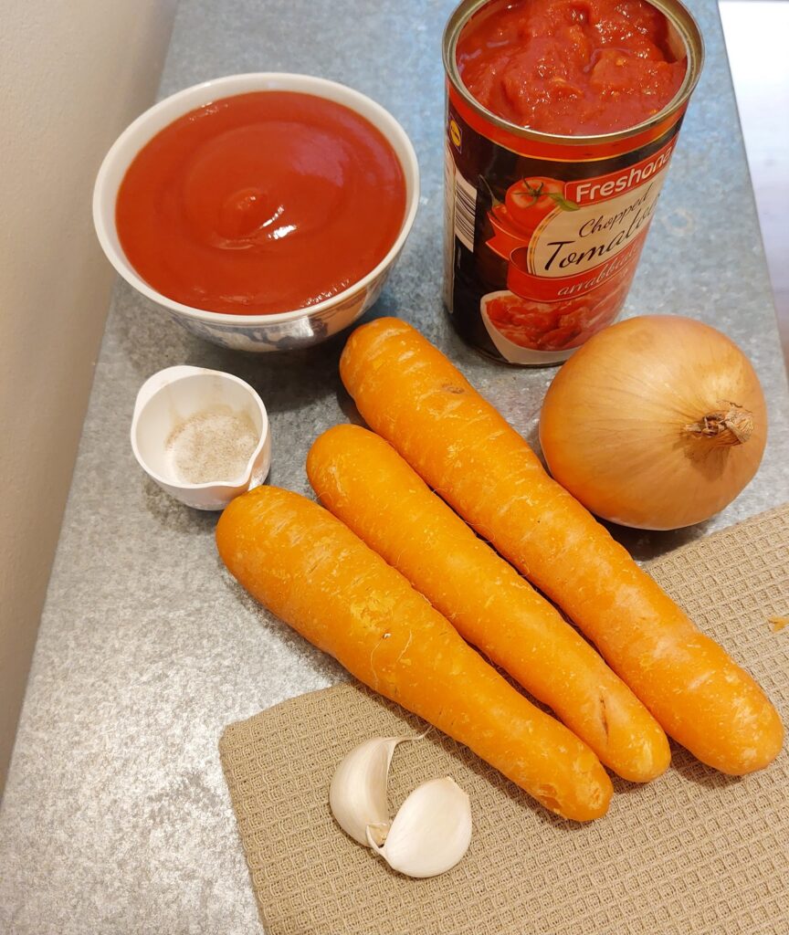 On a work bench the ingredients of the super simple tomato soup are laid out: passata, canned crushed tomatoes, one onion, three carrots, two cloves of garlic, a mixture of salt and pepper in a white cup.