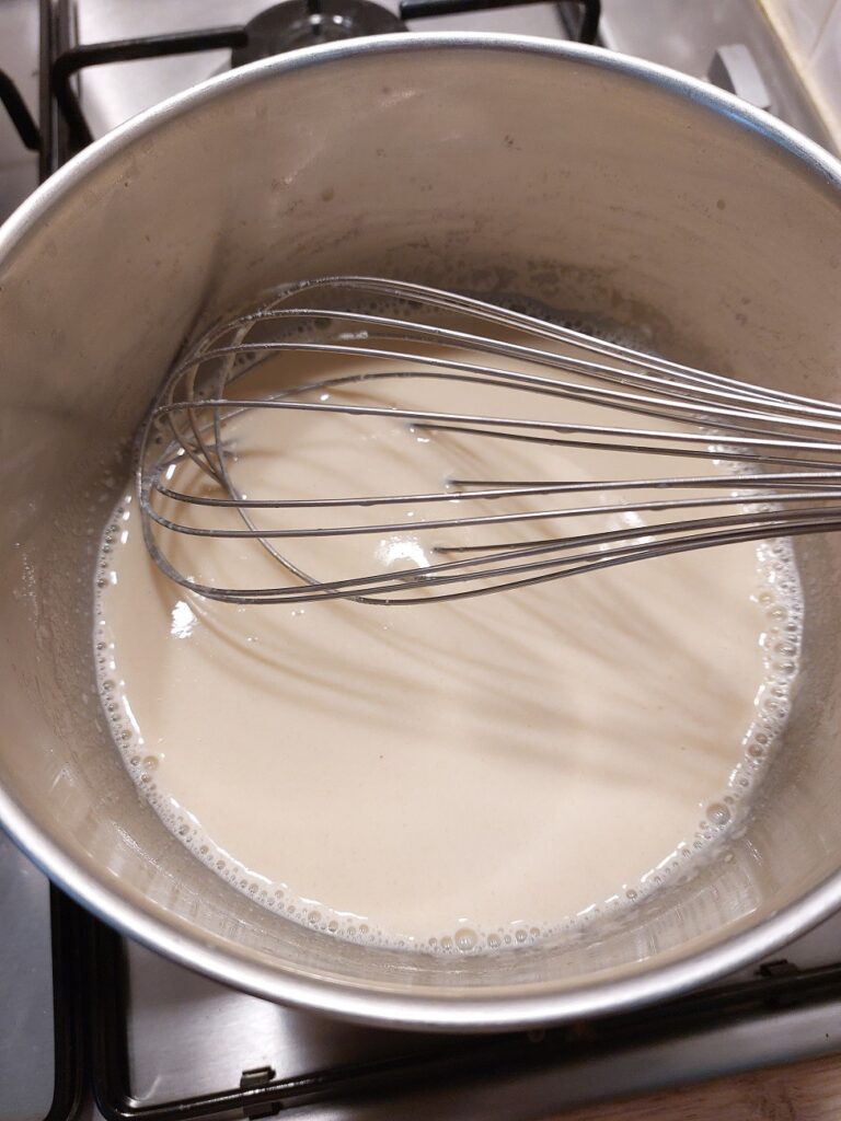 A white sauce being whisked in a silver pot, before adding the cheese to a mac n cheese