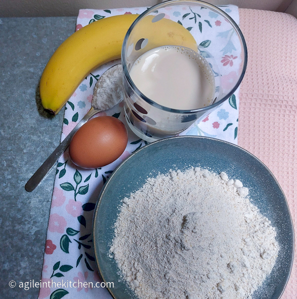 banana waffle ingredients laid out on a pink and flowery background: one banana, a glass of milk, one egg, a teaspoon of baking powder and a plate with oat flour