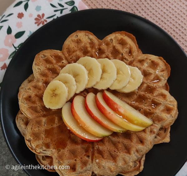 Two banana waffles layered on a dark grey plate, topped with banana and apple slices, drizzled with honey.
