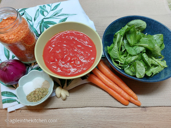 Ingredients for healthy tomato sauce are laid out on a kitchen towel with a printed green lead pattern. One onion, one glass bottle of red lentils, a green bowl of crushed tomatoes, spices in a bowl: oregano, salt, white pepper and sugar, three cloves of garlic, four carrots, a blue bowl of fresh spinach.