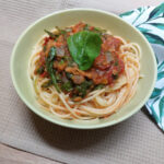 A green bowl filled with spaghetti and healthy tomato sauce, topped with a green leaf of fresh spinach. In the background is a white kitchen towel printed with green leaves.