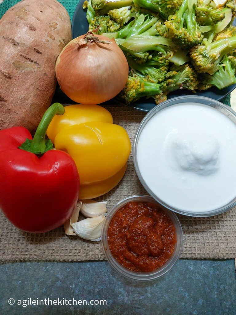 Sweet potato curry ingredients laid out. One sweetpotato, two halves of red and yellow pepper, one onion, a plate of broccoli, a bowl of coconut milk, a smaller bowl of red curry paste, three cloves of garlic