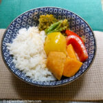 Sweet potato curry plated in a blue patterned bowl with white basmati rice on one side and broccoli, red and yellow peppers and sweet potato on the opposite side. Sitting on a green and beige table cloth.