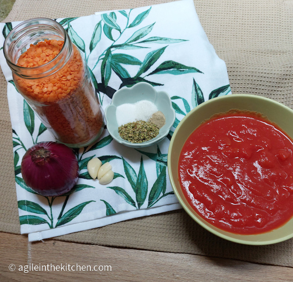 Ingredients to tomato sauce lined up on a kitchen towel with green leaves. One red onion, a glass bottle with red lentils, three garlic cloves, a bowl with oregano, salt, sugar and white pepper, a green bowl with crushed tomatoes. 