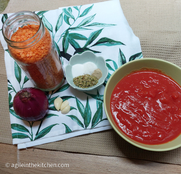 Ingredients for the best adaptable basic tomato sauce laid out. One onion, a glass jar with red lentils, three cloves of garlic, a bowl with oregano, salt, sugar and white pepper, a green bowl with crushed tomatoes. All sitting on top of a kitchen towel printed with green leaves.