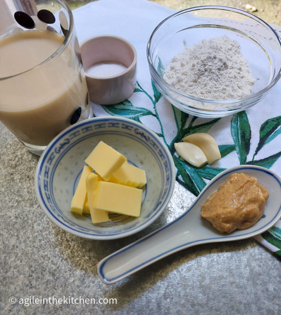 Béchamel and Dijon mustard sauce ingredients laid out, a glass of oat milk, a small pink cup of salt, a glass bowl of flour, two cloves of garlic, a small cup of butter cut up in smaller pieces, and a ceramic spoon of Dijon mustard.