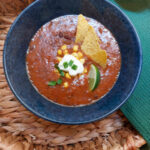 A braided basket with a green cloth, on top there is a blue bowl with black bean soup topped with nachos, corn, a wedge of lime, a dollop of creme fraiche and some green onions