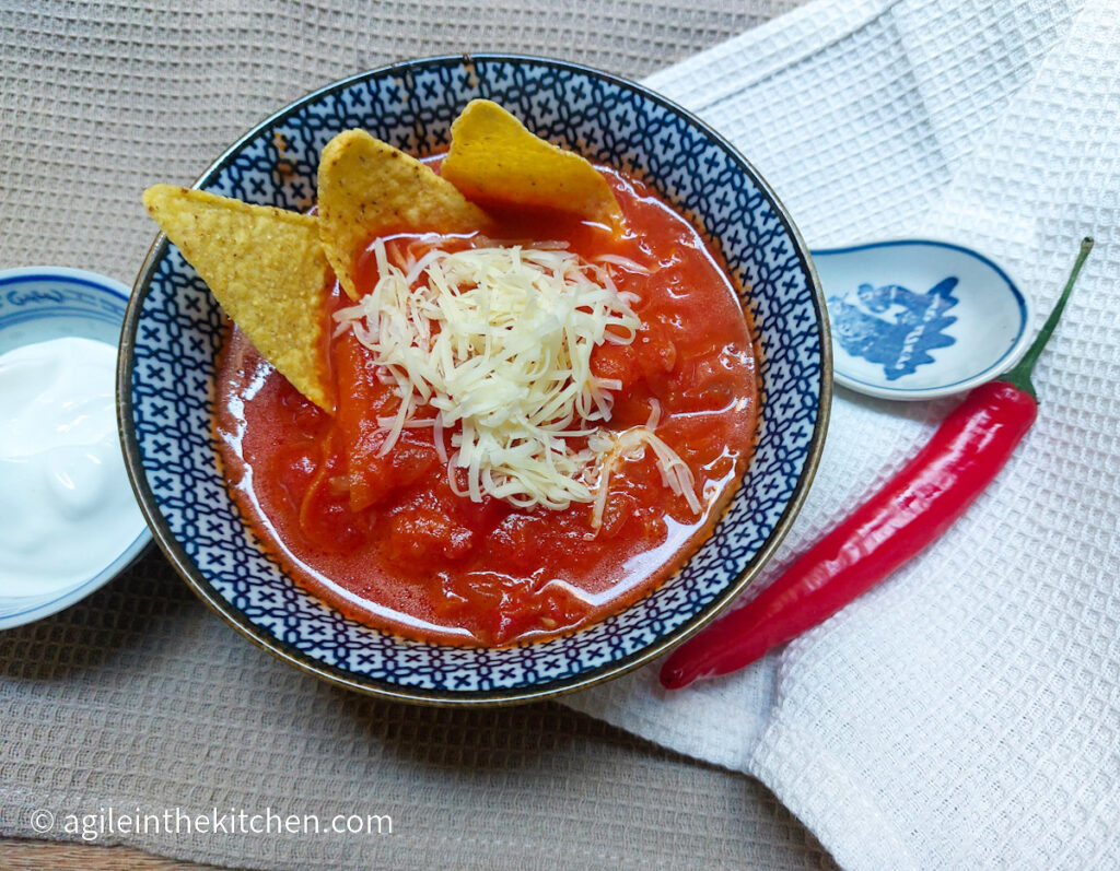 Spicy tomato soup in a blue patterned bowl, with nacho chips, shredded cheese and sliced chilies. Next to a smaller bowl of Greek yogurt with a red chili pepper on the other side.
