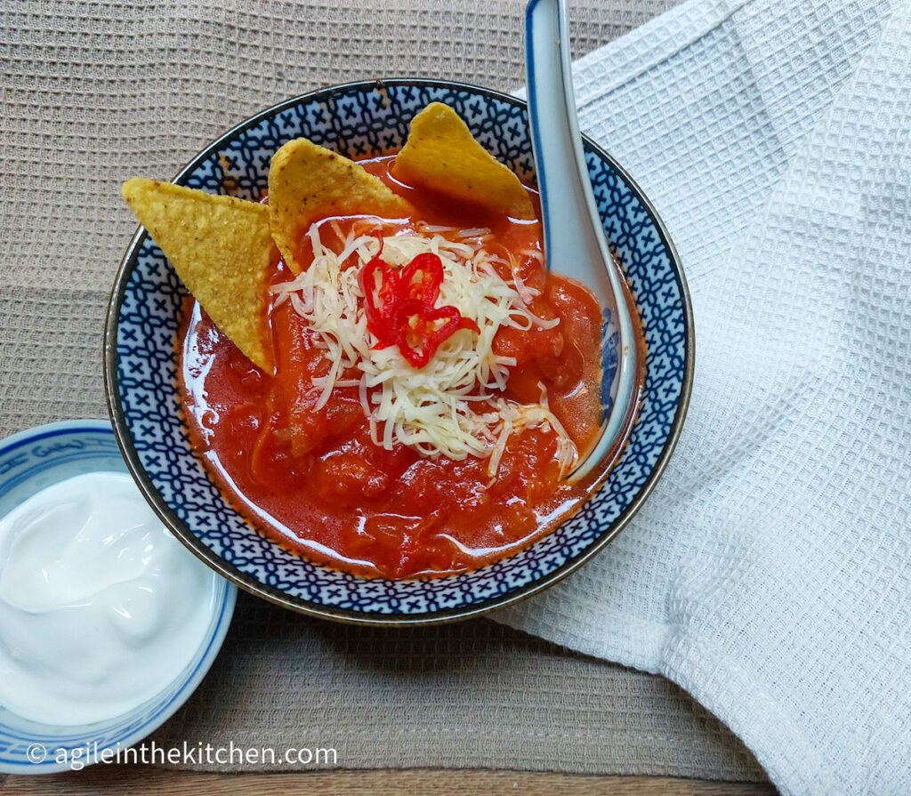 Spicy tomato soup in a blue patterned bowl, with nacho chips, shredded cheese and sliced chilies. Next to a smaller bowl of Greek yogurt.