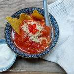 Spicy tomato soup in a blue patterned bowl, with nacho chips, shredded cheese and sliced chilies. Next to a smaller bowl of Greek yogurt.