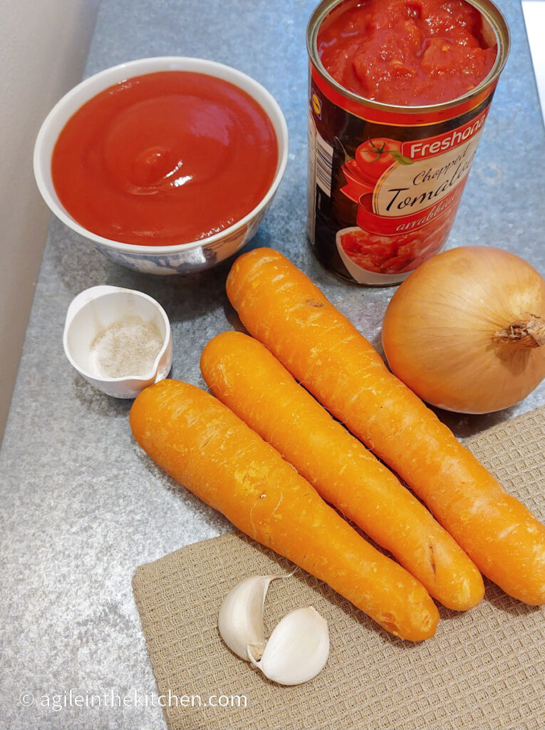 On a work bench the ingredients of the super simple tomato soup are laid out: passata, canned crushed tomatoes, one onion, three carrots, two cloves of garlic, a mixture of salt and pepper in a white cup.