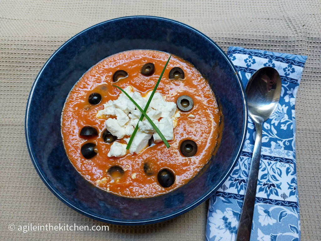 Mediterranean soup in a blue bowl, topped with crumbled feta cheese, black olives and green onions, next to a folded blue napkin with a silver spoon on top of it.