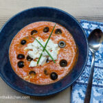 Mediterranean soup in a blue bowl, topped with crumbled feta cheese, black olives and green onions, next to a folded blue napkin with a silver spoon on top of it.