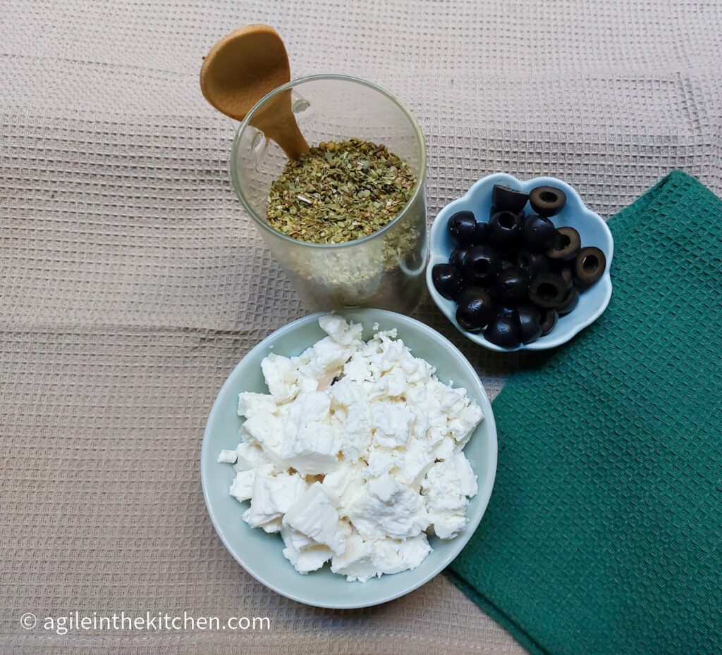 Ingredients to adapt plain tomato soup into a Mediterranean tomato soup laid out. Oregano in a glass bottle, cut up black olives in a small bowl and crumbled feta cheese in a larger bowl.