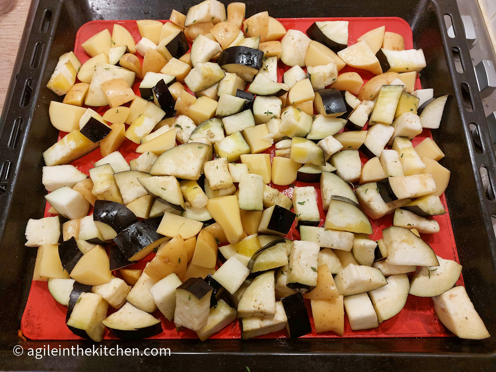 First layer of roasted aubergine mix ready to bake in the oven with diced aubergine and diced potatoes on a red silicone baking sheet