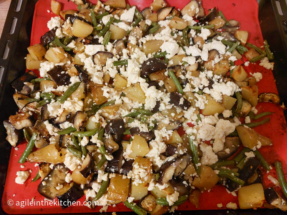 Roasted aubergine mix ready to bake in the oven with diced aubergine, diced potatoes, cut up green beans and crumbled feta cheese on a red silicone baking sheet.