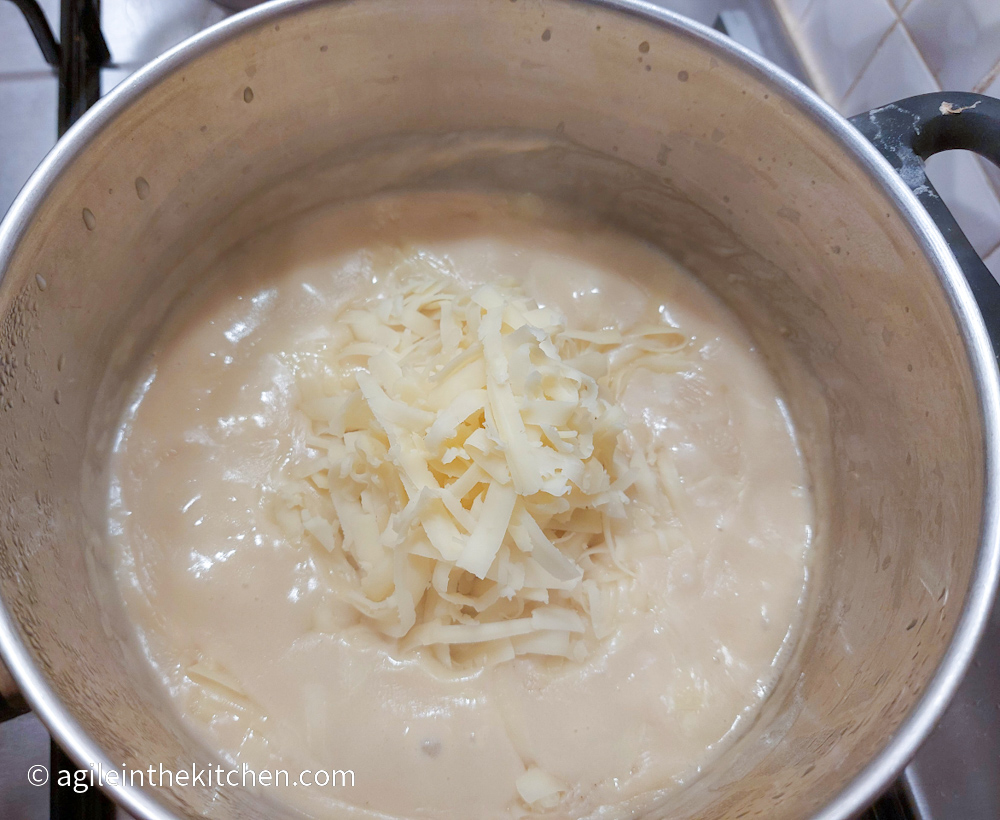 Adding shredded cheese to the bechamel sauce, cooking in a silver coloured pot on a gas stove top.