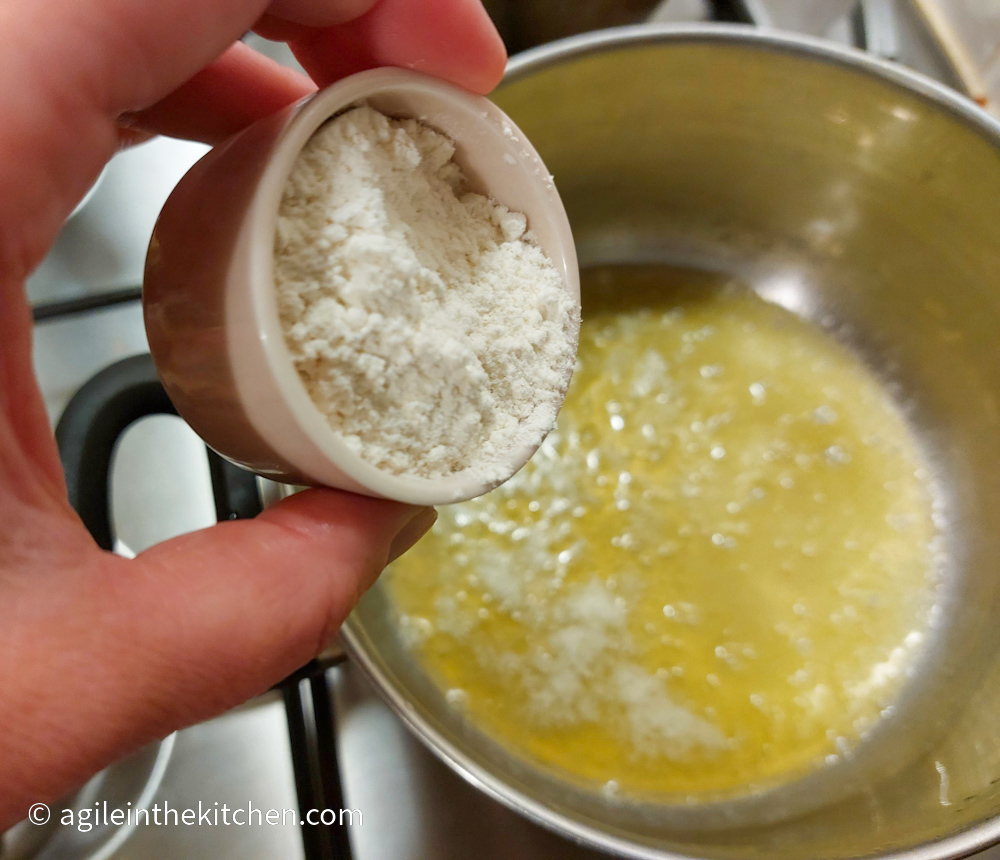 A hand adds flour from a pink pot to melted butter in a pot on a stove top to create a basic roux.