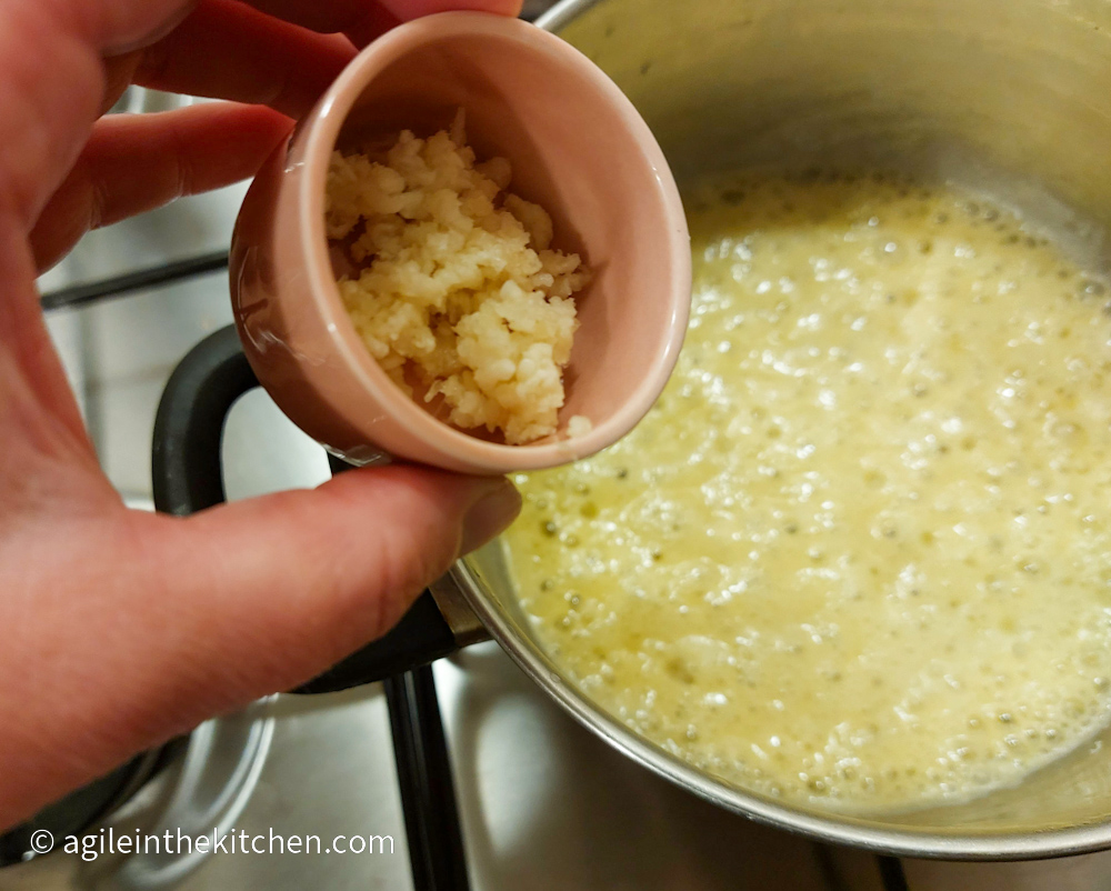 Basic roux cooking on the stove, while a hand adds minced garlic from a pink bowl.