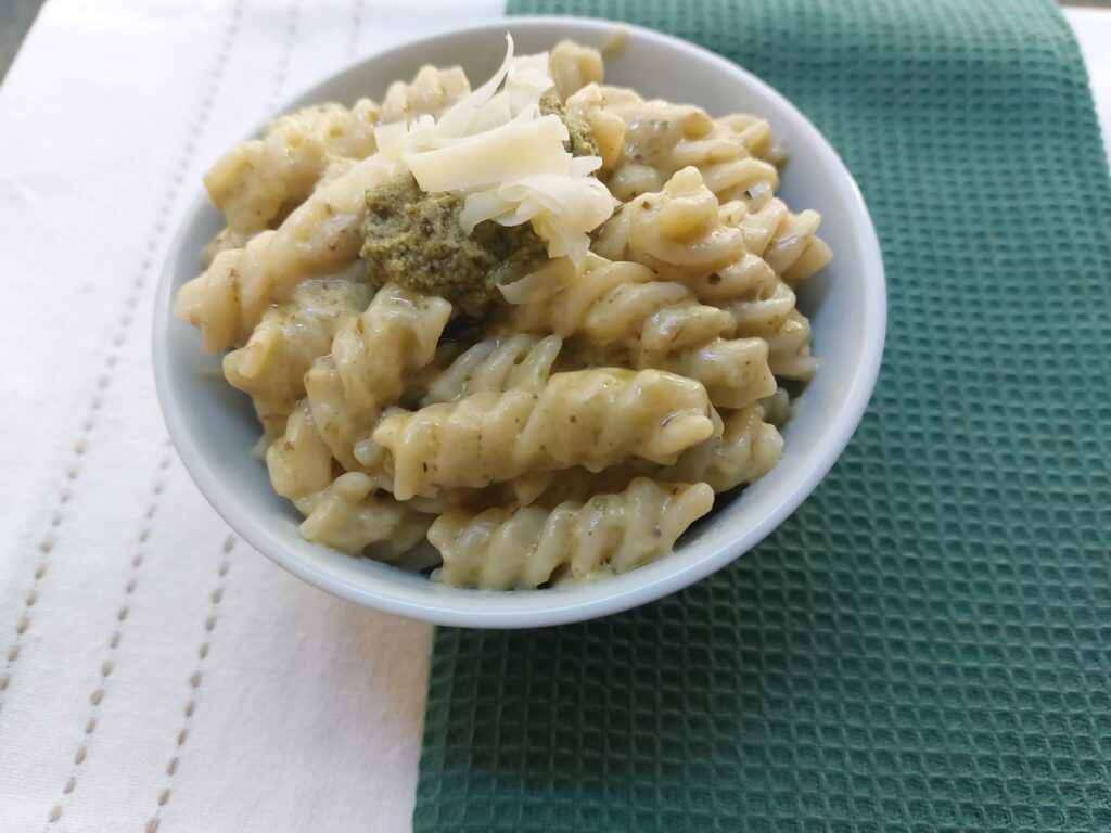 A bowl of green Mac n' Cheese topped with green pesto and shredded cheese, sitting on top of a green and white table cloth.