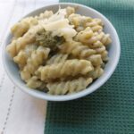 A bowl of green Mac n' Cheese topped with green pesto and shredded cheese, sitting on top of a green and white table cloth.