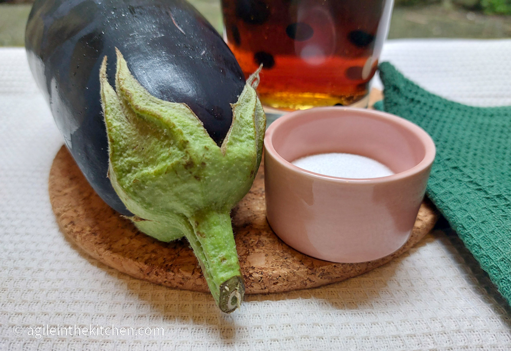 A close up of an aubergine, a glass of red ale and a pot of salt and a green napkin, resting on a cork cutting board