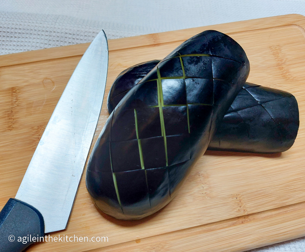 An aubergine cut in half then scored, laying on a wooden cutting board with a knife