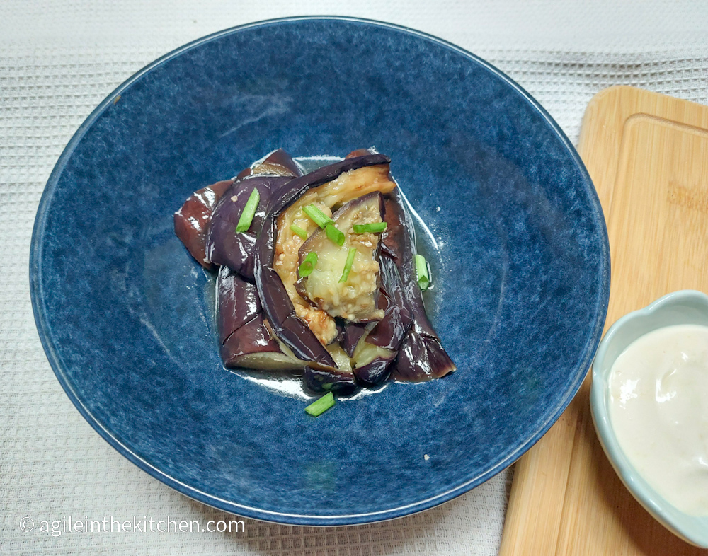 Red Ale aubergine cooked and plated in a blue bowl, sprinkled with green onions, next to a wooden cutting board and dressing in a bowl