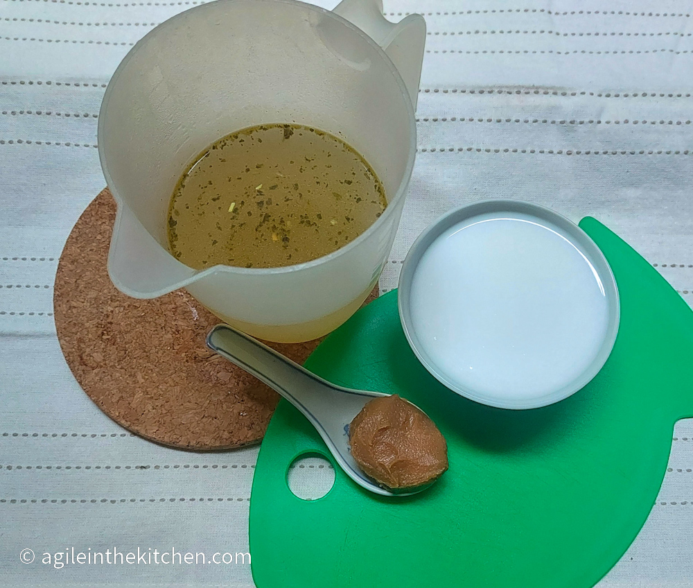 Ingredients to make the Coconut Ramen broth laid out on a white table cloth, a brown pot holder and a green cutting board in the shape of a fish. Vegetable broth in a liter measuring cup, miso paste on a spoon and coconut milk in a small bowl.