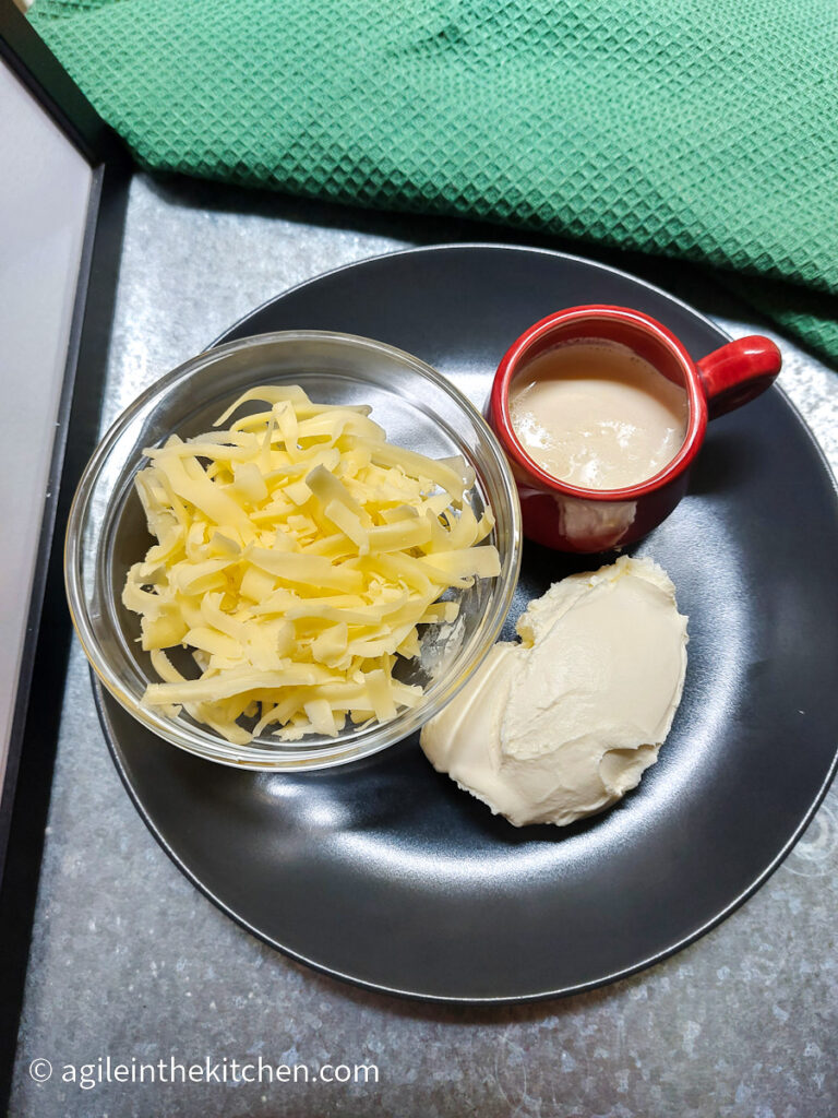 Creamy Mac n' Cheese ingredients plated on a black plate, sitting on a silver background with a green cloth napkin. Ingredients: Shredded cheese, cream cheese and heavy cream