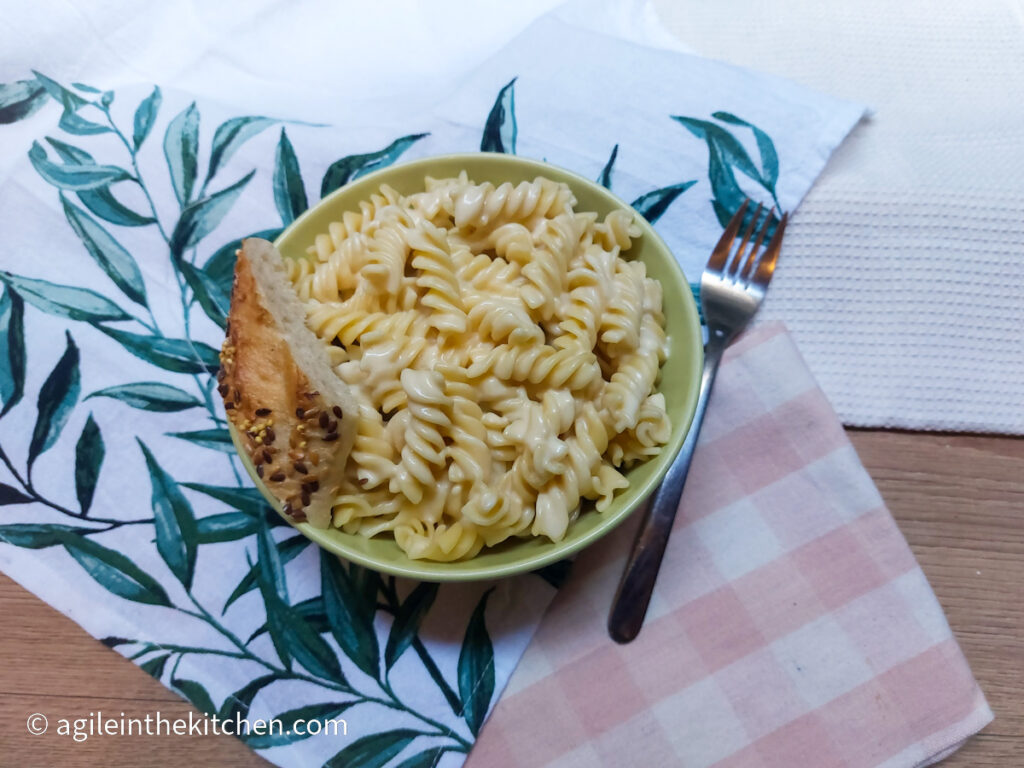 Creamy Mac n' Cheese in a green bowl, with a piece of bread on the side and a fork. Placed on two table cloths, one pink gingham and one with green leaves.